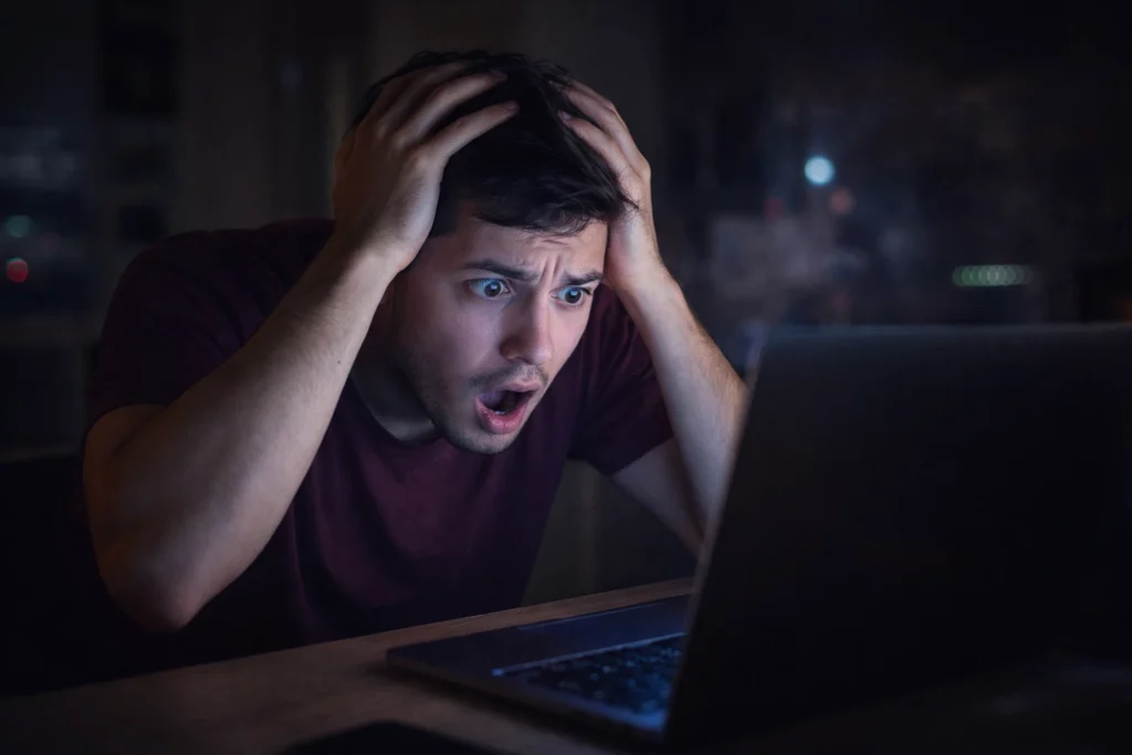Young man holding his head in shock while looking at a computer screen after discovering malware